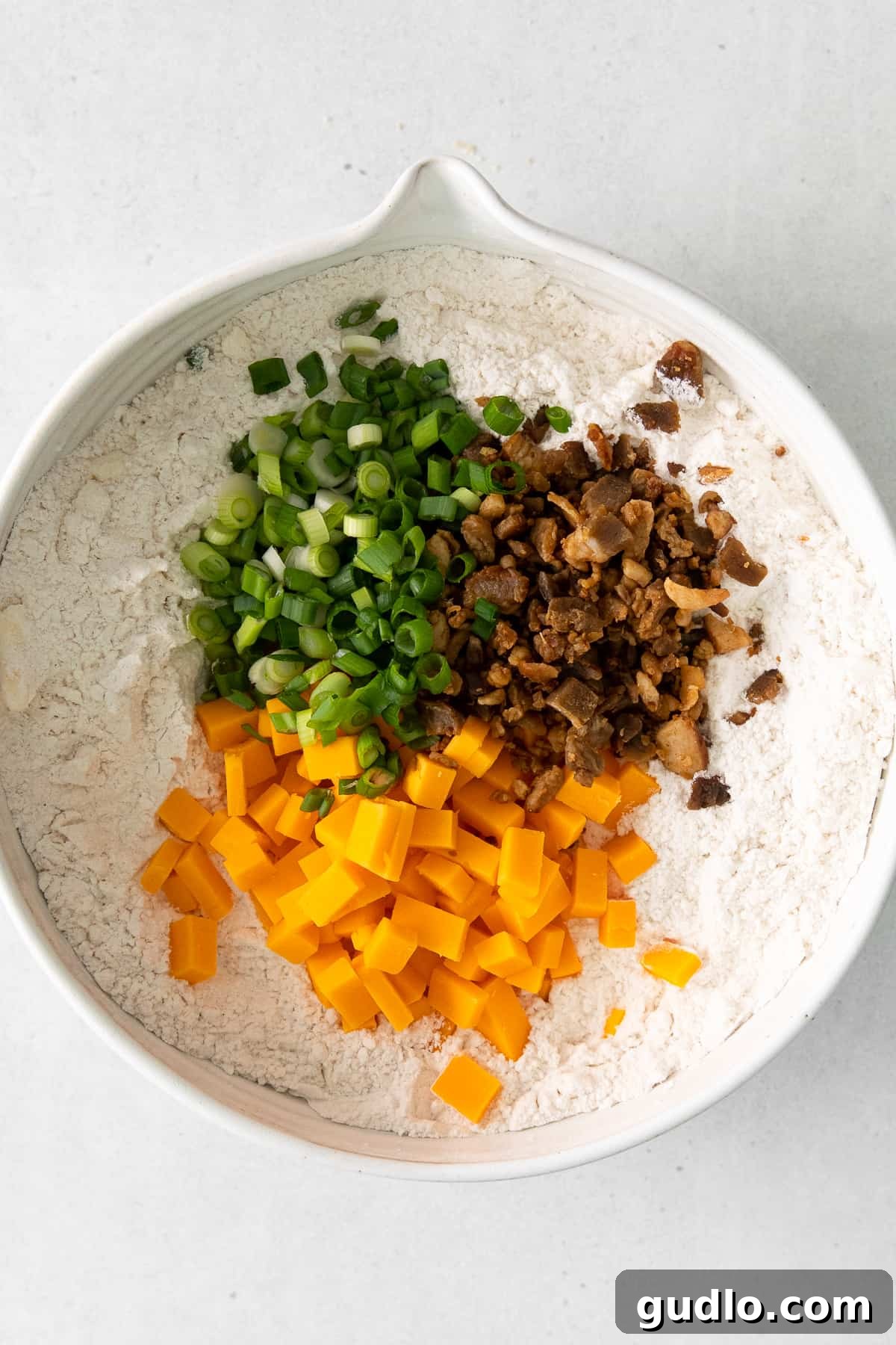 Essential Ingredients for Savory Scones A vibrant collection of savory scone ingredients in a bowl, featuring freshly grated cheddar cheese, crispy bacon bits, and chopped chives nestled among the dry flour mixture.