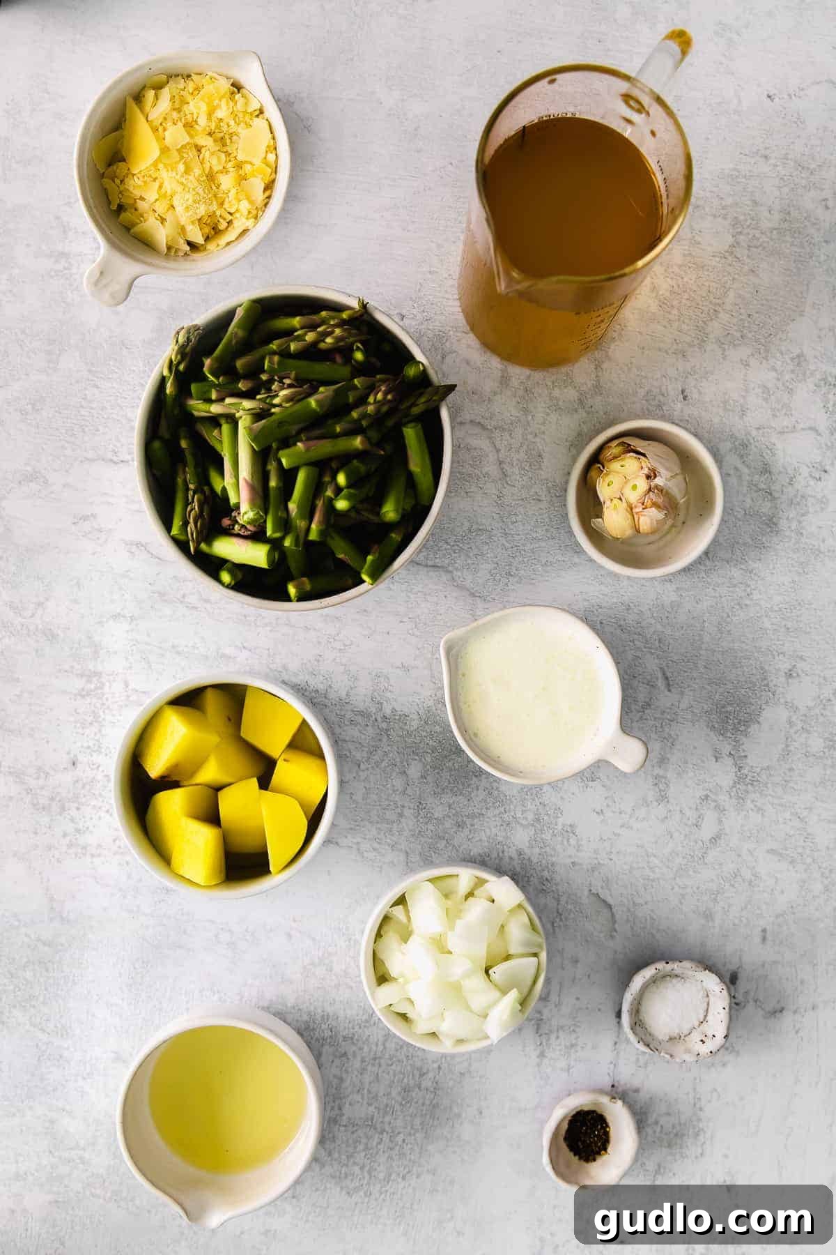 Assortment of fresh ingredients for asparagus soup laid out on a kitchen counter: asparagus, onion, potato, garlic, and a bottle of olive oil.