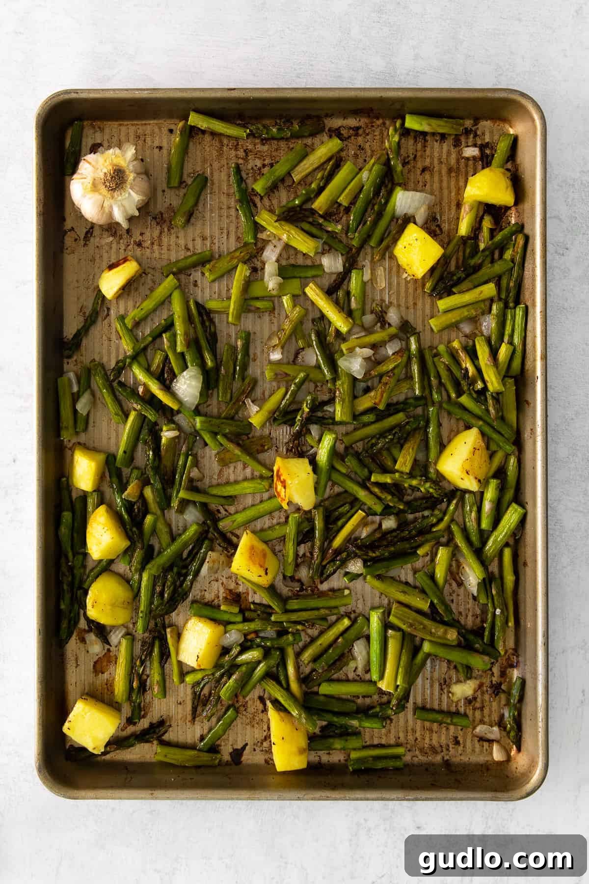 Sheet pan with roasted asparagus, chopped onion, potato, and a head of garlic, seasoned with salt, pepper, and olive oil.