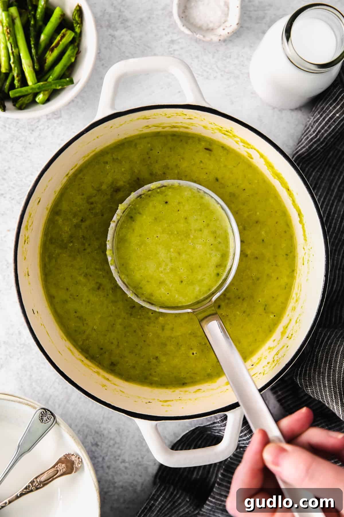 A ladle of creamy asparagus soup being served from a large pot.