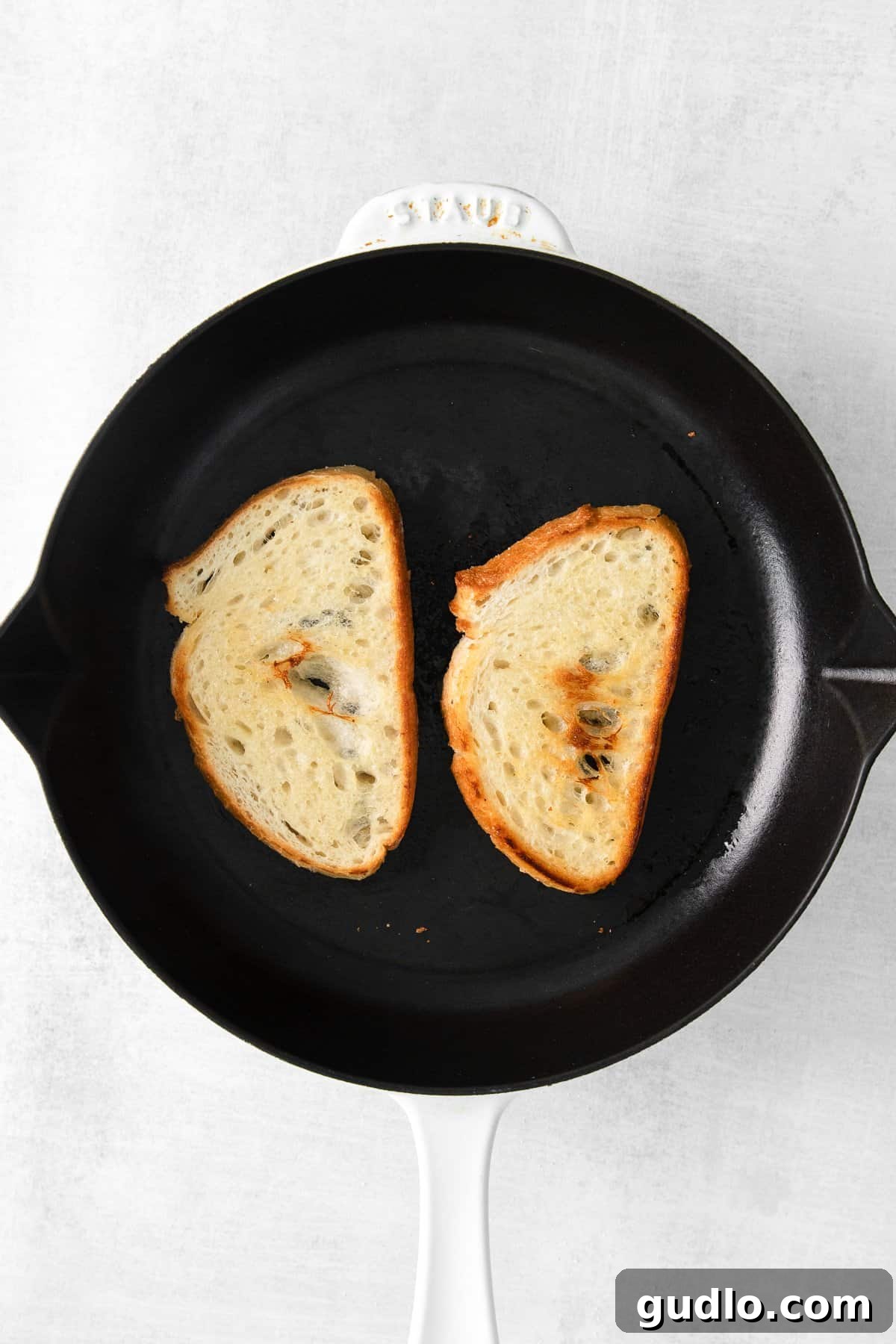 Two thick slices of sourdough bread sizzling in a seasoned cast iron skillet, turning golden brown as they toast, preparing them for the creamy mascarpone spread.