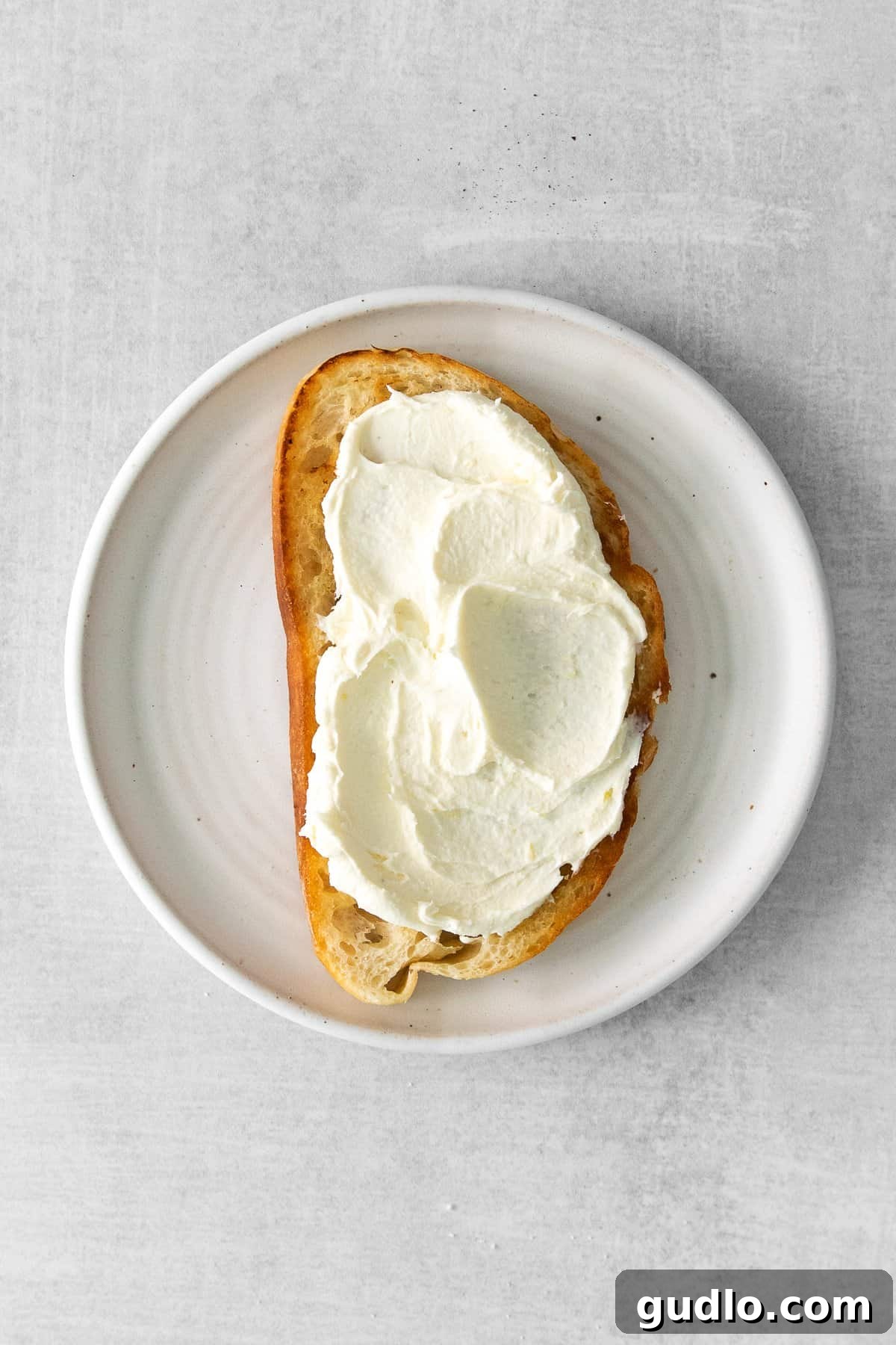 A single slice of perfectly toasted sourdough bread, generously spread with a thick layer of creamy white mascarpone cheese, ready for toppings.
