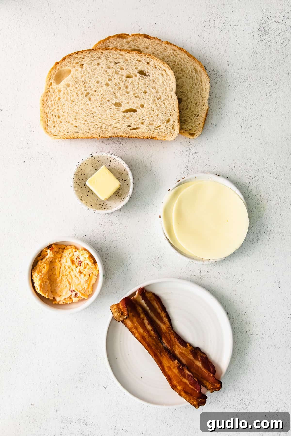 A collection of fresh ingredients laid out on a clean countertop, including bread, pimento cheese, smoked gouda, bacon, and butter, ready for assembly.