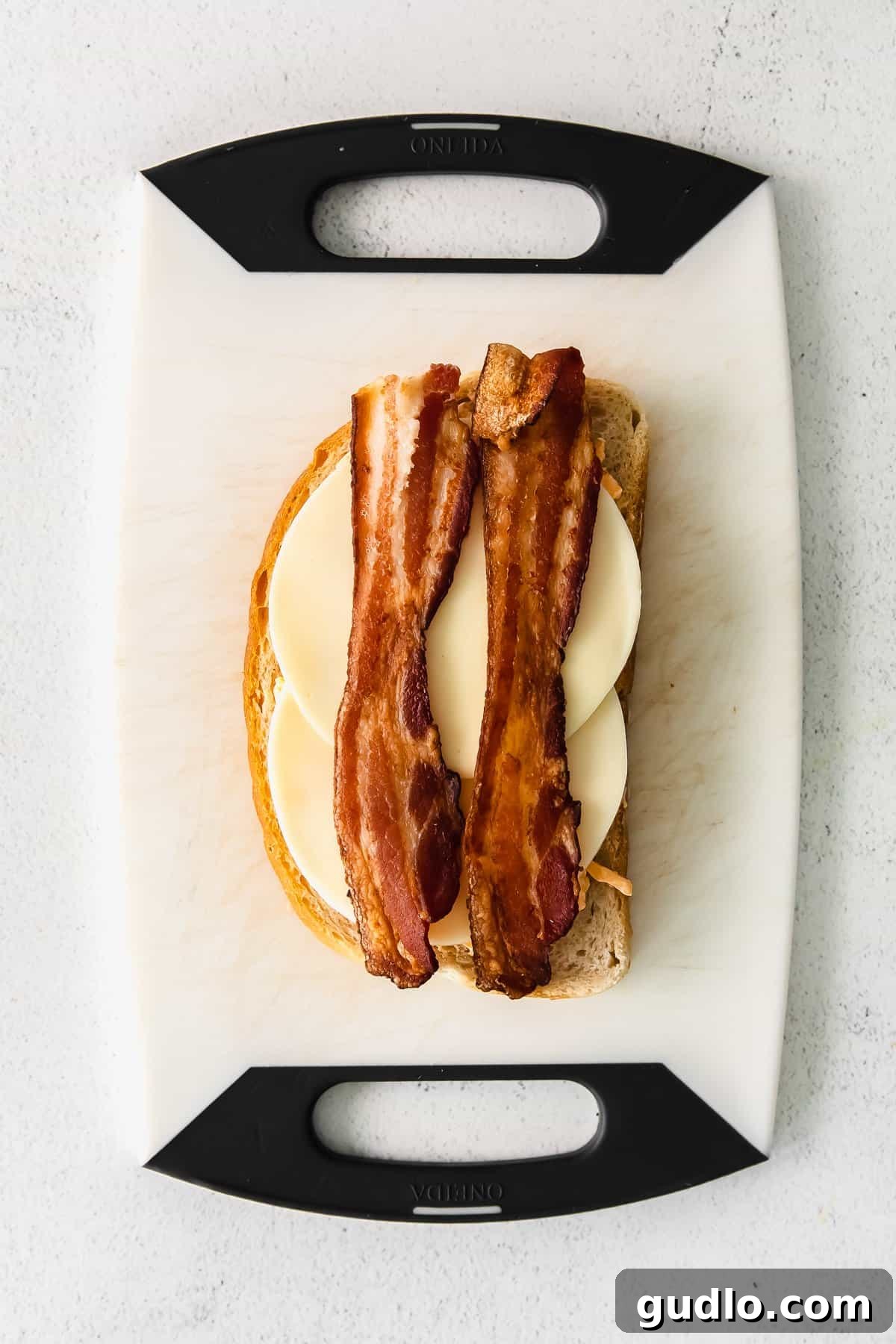 An open-faced grilled cheese showing layers of melted pimento cheese, smoked gouda, and bacon, ready to be topped with the second slice of bread.
