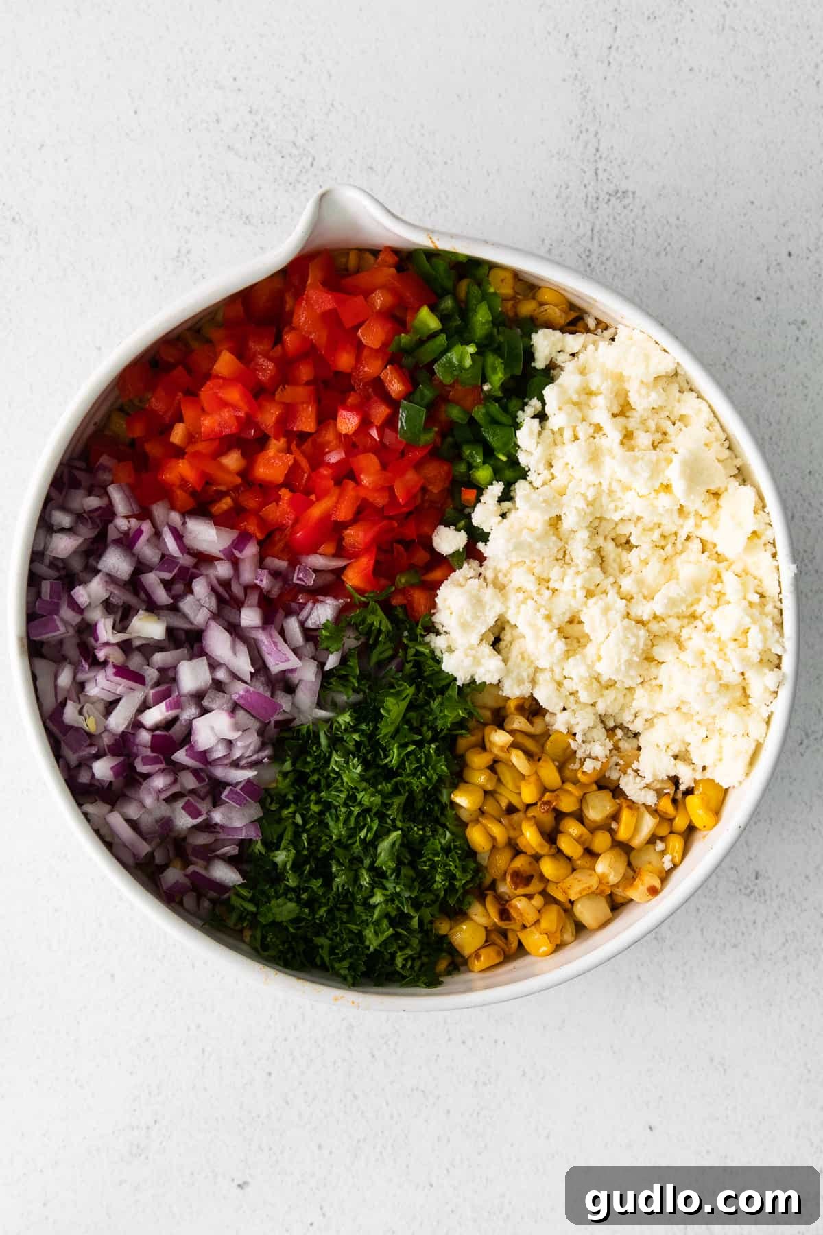A large white bowl filled with the colorful ingredients for Mexican corn salad, showcasing the vibrant corn, peppers, and onions.