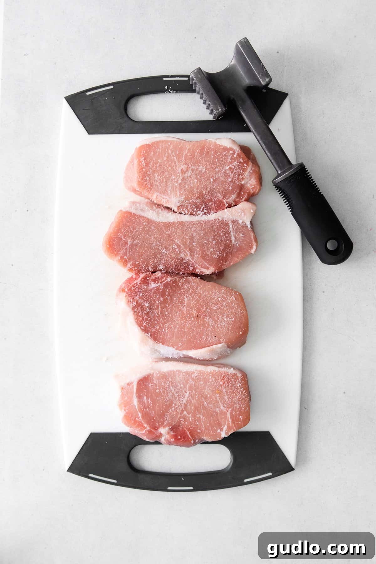 Golden Parmesan Pork Chops 4 Pork chops being pounded thin on a cutting board, covered with plastic wrap.