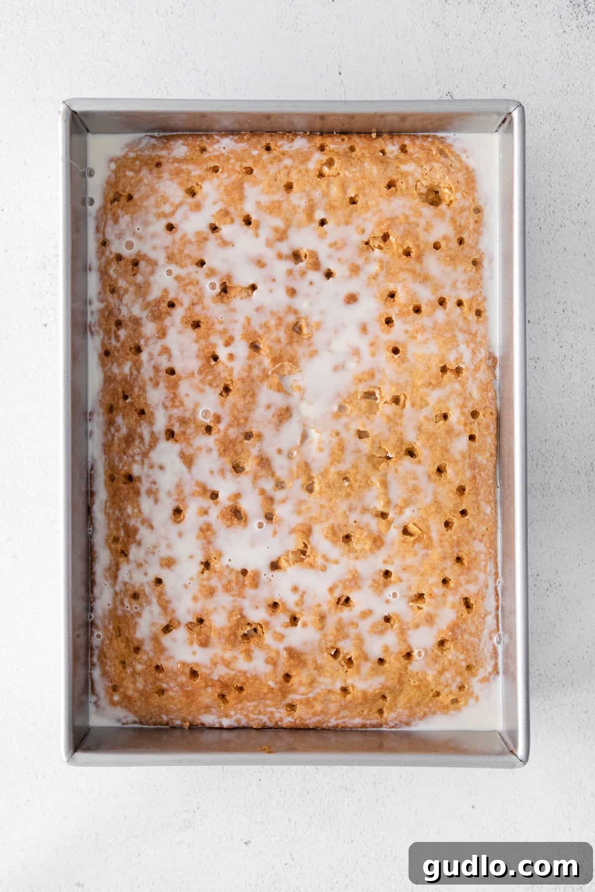 Tres Leches milk mixture being poured over a freshly baked sponge cake, soaking into the poked holes.