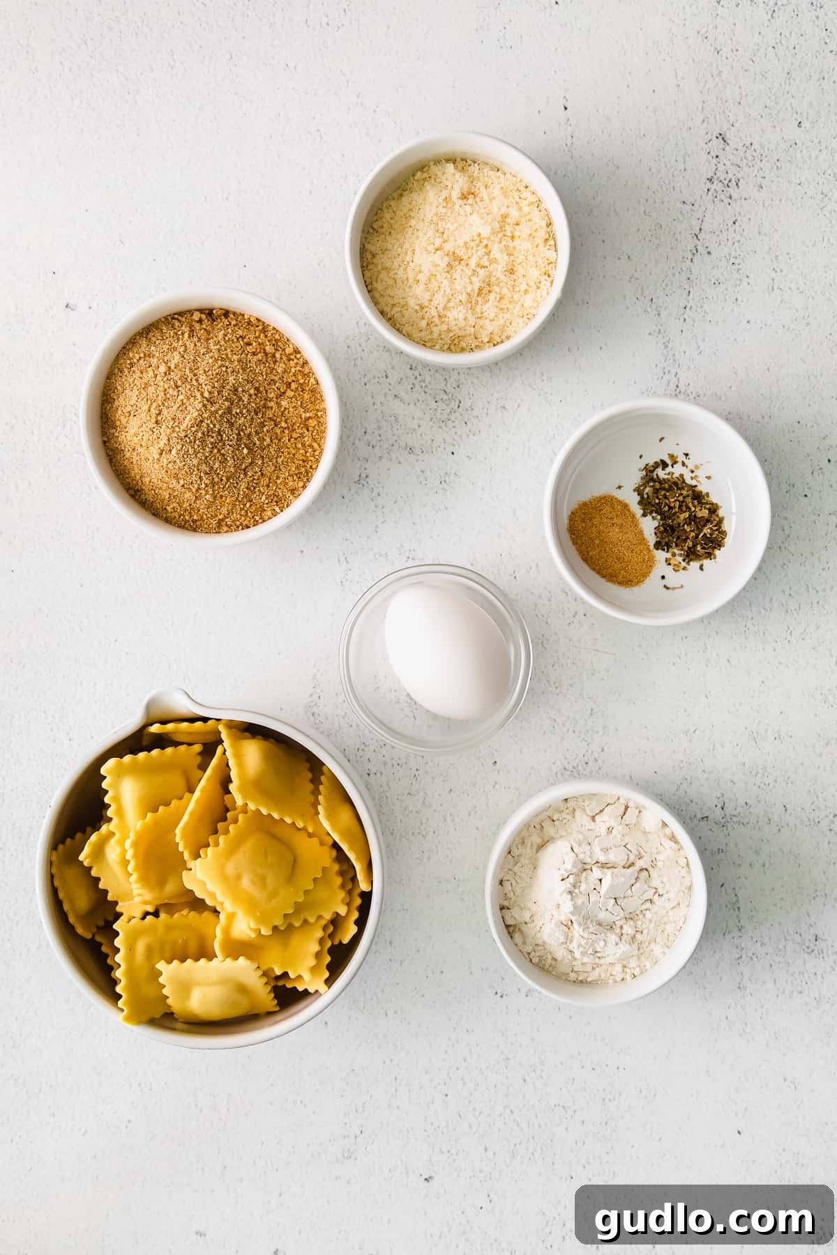 A close-up shot of small bowls containing the key ingredients for air fryer ravioli: fresh ravioli, all-purpose flour, a beaten egg, a mixture of breadcrumbs and Parmesan cheese, and a blend of garlic powder and Italian seasoning.
