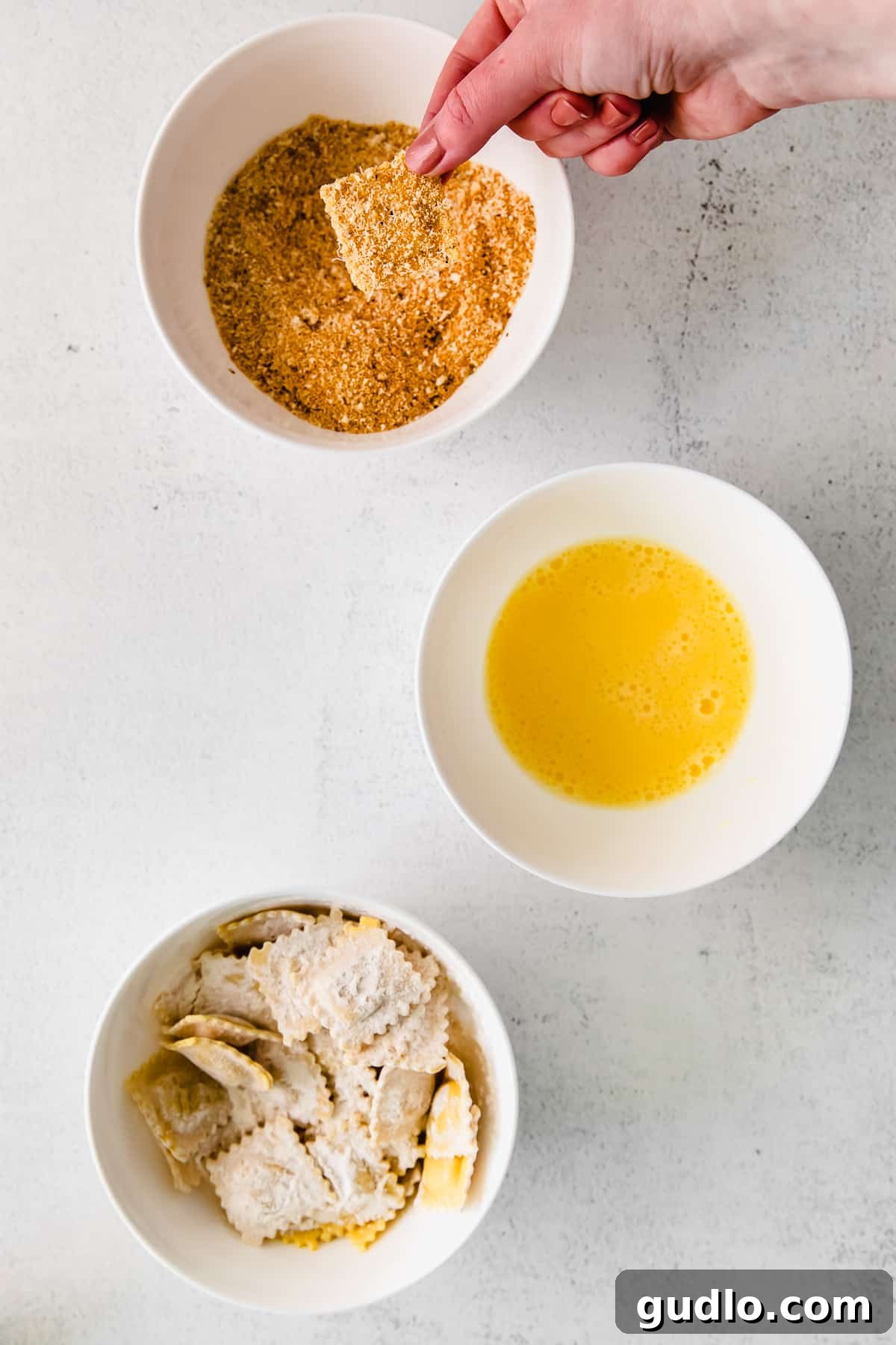 A hand gently dips a flour-coated ravioli into a bowl filled with a golden breadcrumb and finely grated Parmesan cheese mixture, ensuring a complete and even coating.