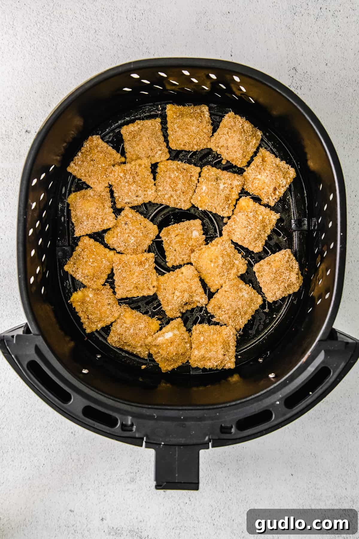 Several breaded ravioli pieces are neatly arranged in a single layer inside an air fryer basket, perfectly spaced for optimal air circulation and even cooking.