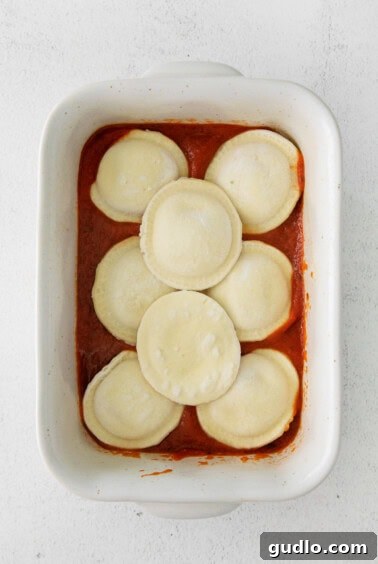 First layer of frozen ravioli on marinara sauce in a white baking dish.