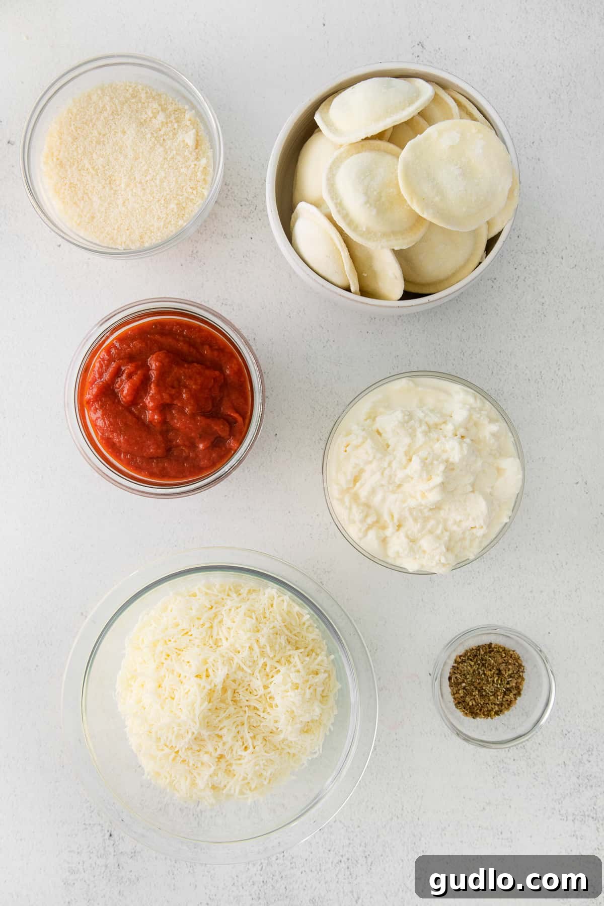 Small bowls containing fresh ingredients for ravioli lasagna: marinara sauce, ricotta cheese, shredded mozzarella, grated parmesan, and a blend of Italian seasonings.