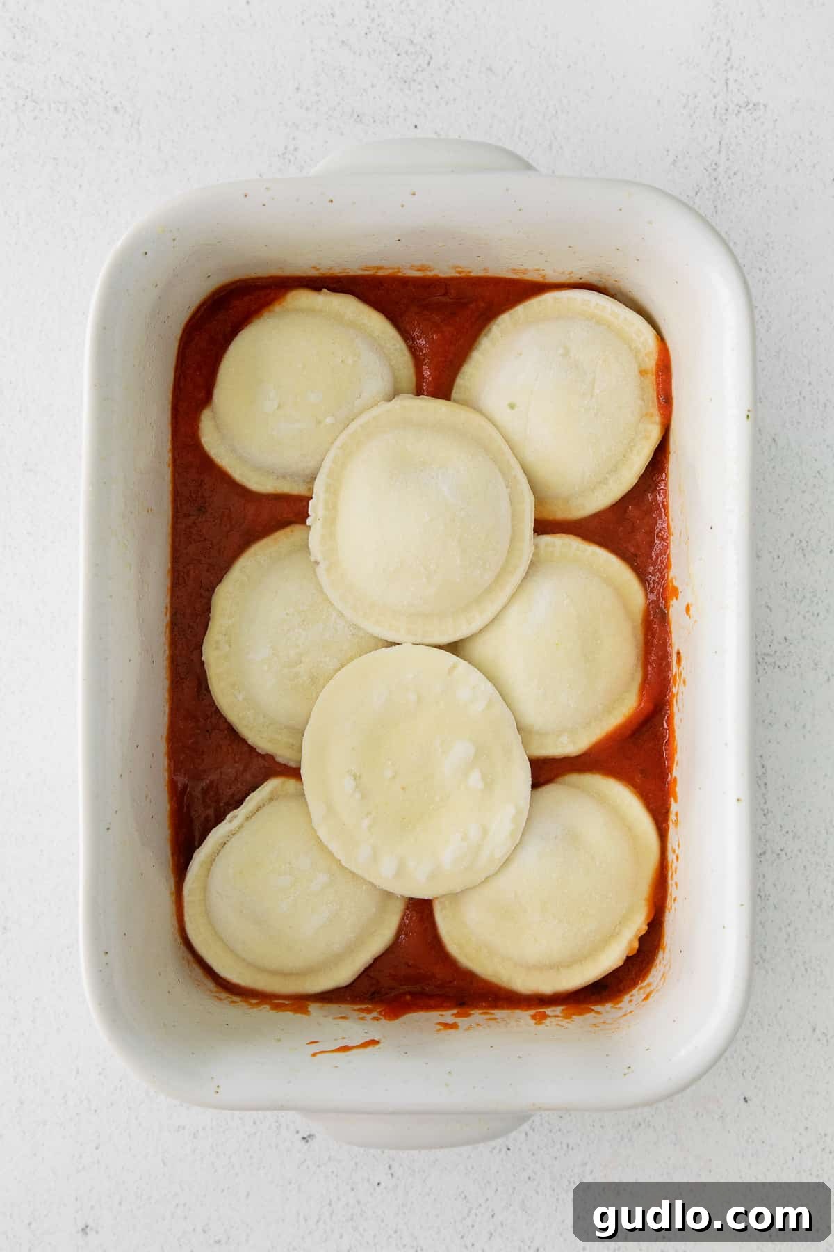 The first layer of frozen ravioli neatly arranged in a casserole dish, partially covered with marinara sauce.
