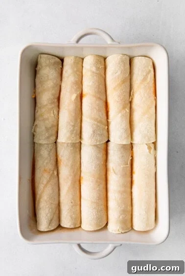 Irresistibly Cheesy Enchiladas 6 Rolled enchiladas neatly arranged in a white baking dish, partially covered in sauce.