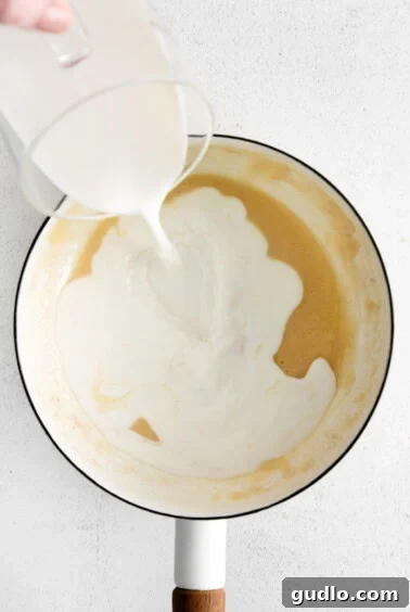 Milk being slowly added to a thickening roux in a pot while being continuously whisked.