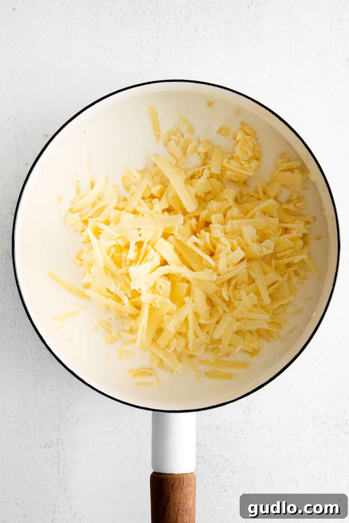Freshly shredded sharp white cheddar cheese being gently stirred into a creamy white sauce in a pot, melting into a luxurious, homogeneous texture.