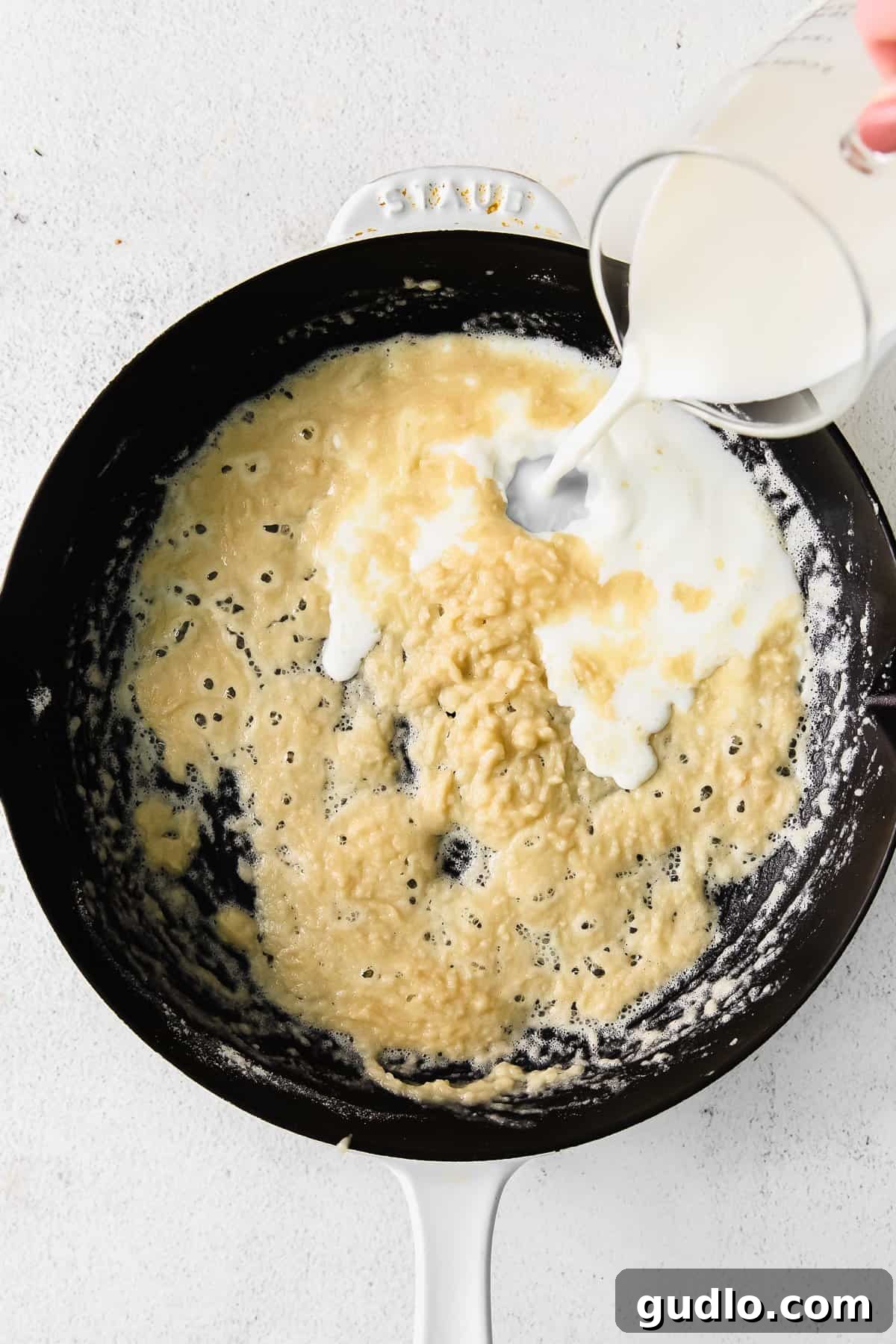 Learn how to make a creamy, white bechamel sauce recipe with just 3 basic ingredients - butter, flour, and milk. A steady stream of milk being poured into a golden roux in a cast iron pan, with a whisk actively combining the ingredients.