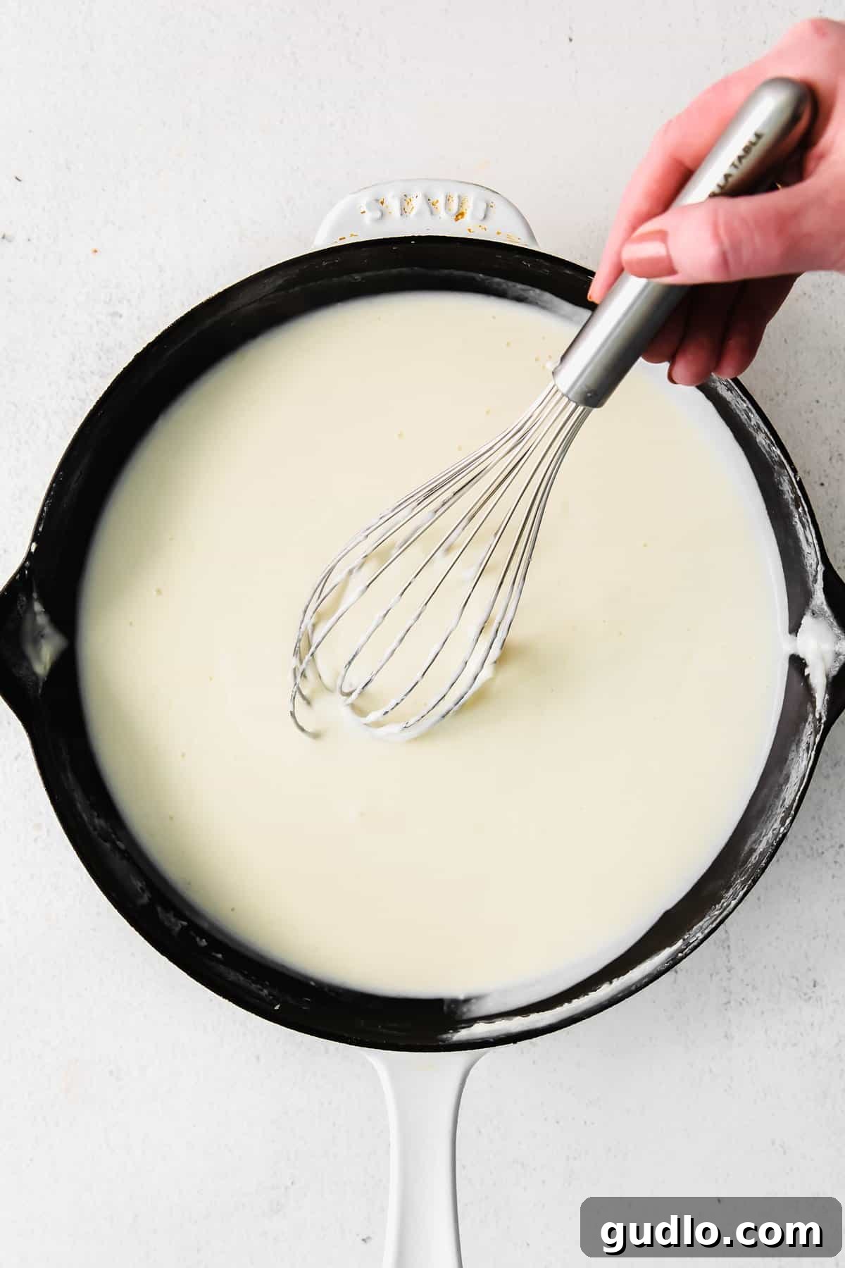 Learn how to make a creamy, white bechamel sauce recipe with just 3 basic ingredients - butter, flour, and milk. A whisk stirring a creamy, white bechamel sauce in a cast iron pan, showing its smooth texture.