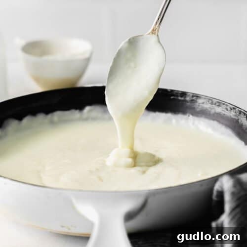 A spoon dripping white sauce into a pan, showcasing the creamy texture of bechamel.