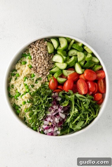 A large white bowl filled with a mix of orzo pasta, green peas, and various fresh vegetables, ready for dressing.