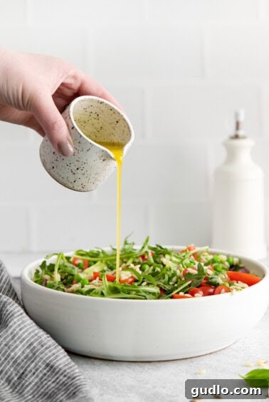 A hand pouring a stream of golden lemon vinaigrette over a mixed salad in a white bowl, ensuring even coating.