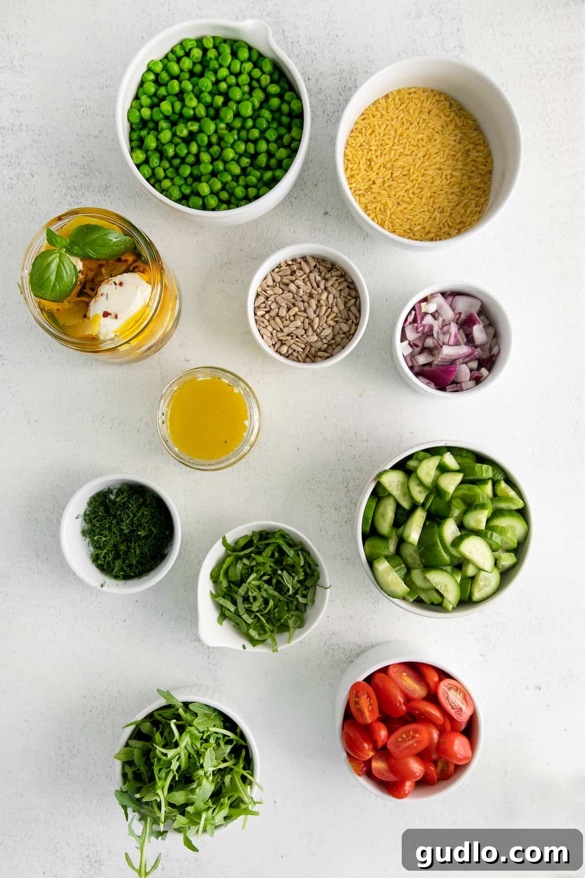 Fresh ingredients for lemon orzo salad, including cucumber, cherry tomatoes, red onion, peas, and fresh herbs, in separate bowls.