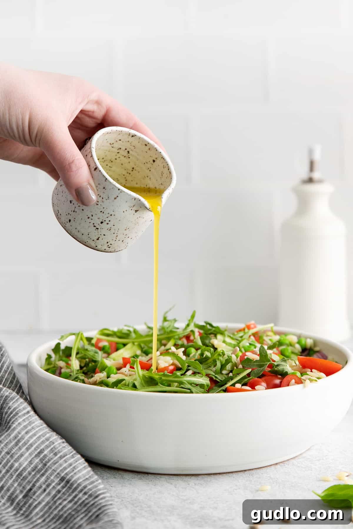 A vibrant stream of lemon vinaigrette being drizzled generously over the mixed orzo salad in a white bowl.