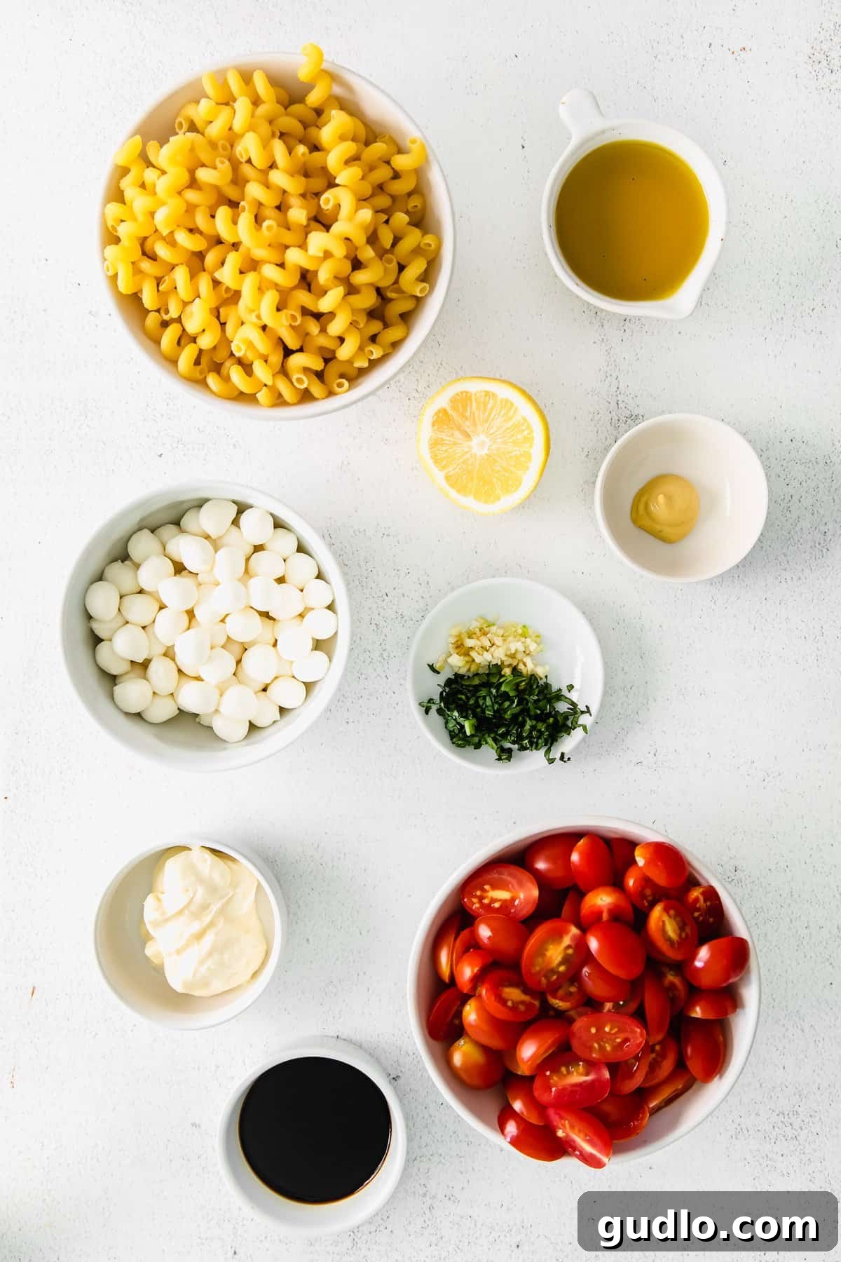 Vibrant Caprese Pasta Toss 3 Small bowls displaying individual ingredients: cavatappi pasta, halved cherry tomatoes, small mozzarella balls, and fresh basil leaves, ready for assembly.