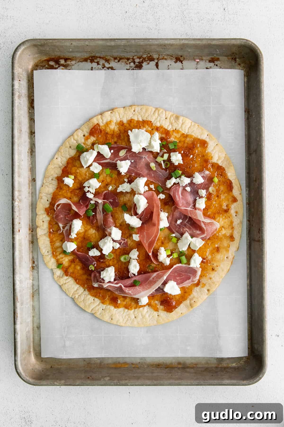 Flatbread Baking in the Oven Fig and goat cheese flatbread baking on a parchment-lined sheet, showing the melted cheese and crispy prosciutto.
