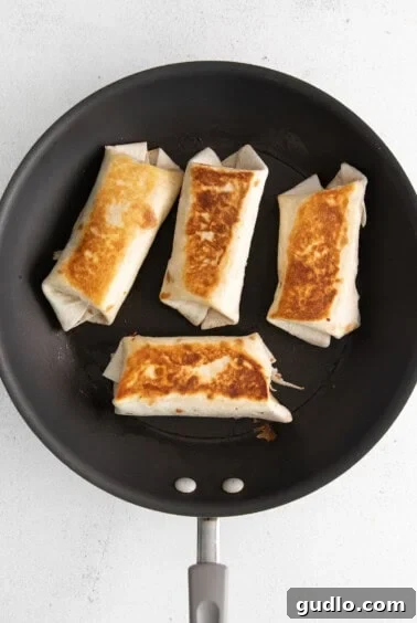 Four burritos searing in a frying pan on a white surface.