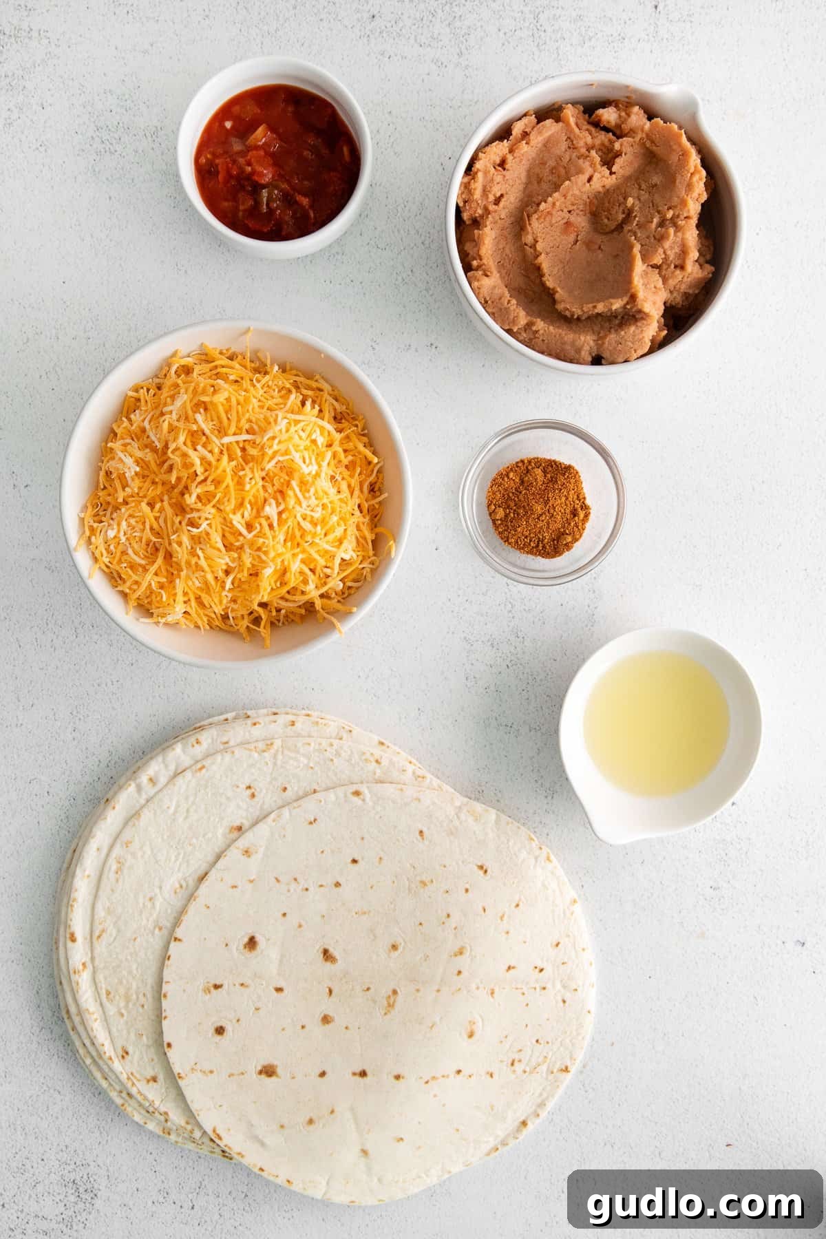 Simple Ingredients for Homemade Bean and Cheese Burritos Various bean and cheese burrito ingredients neatly arranged in small bowls, including tortillas, refried beans, salsa, taco seasoning, and shredded cheese.