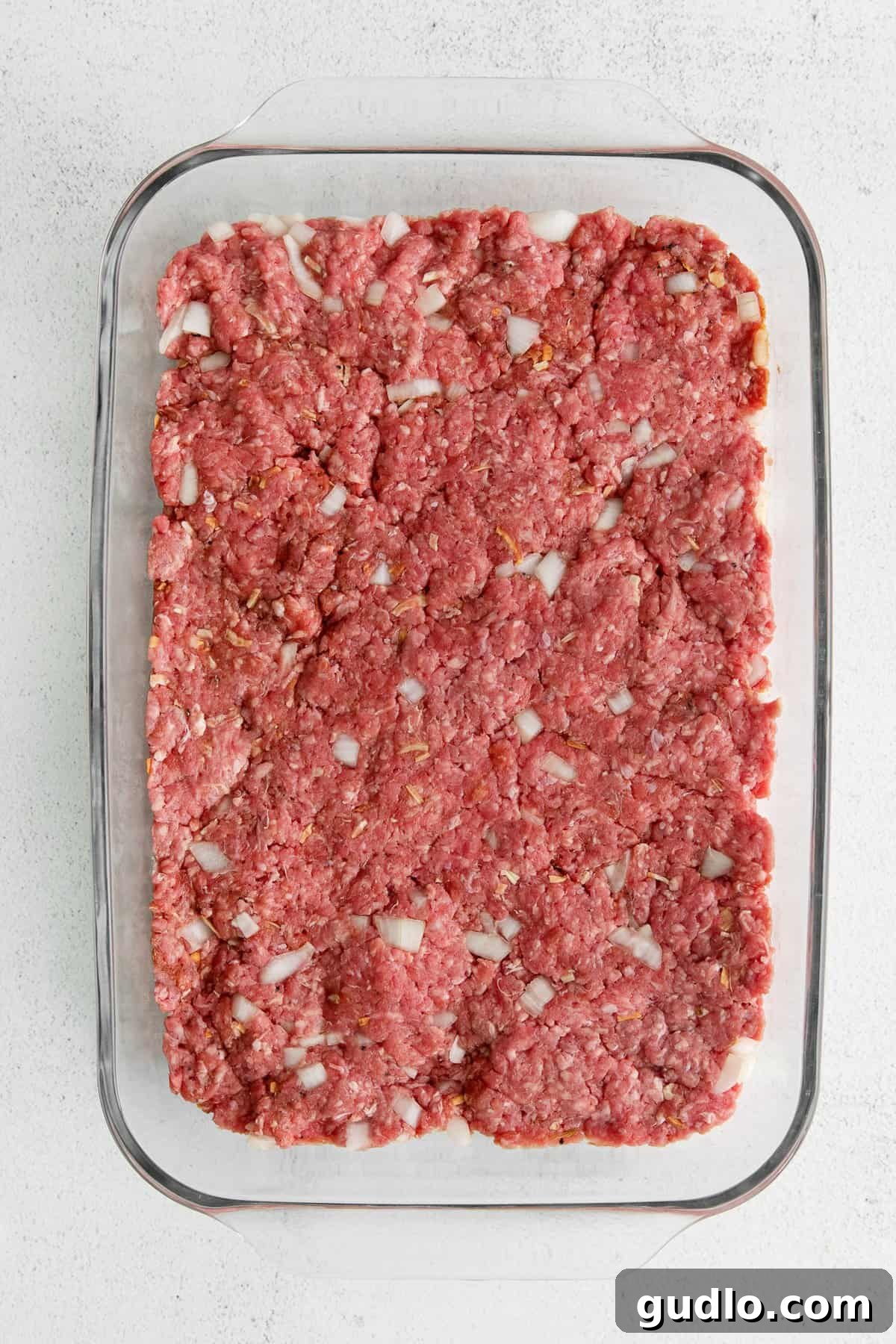 Pre-Bake: Giant Patty for Cheeseburger Sliders Large hamburger patty pressed into a casserole dish, ready for baking.