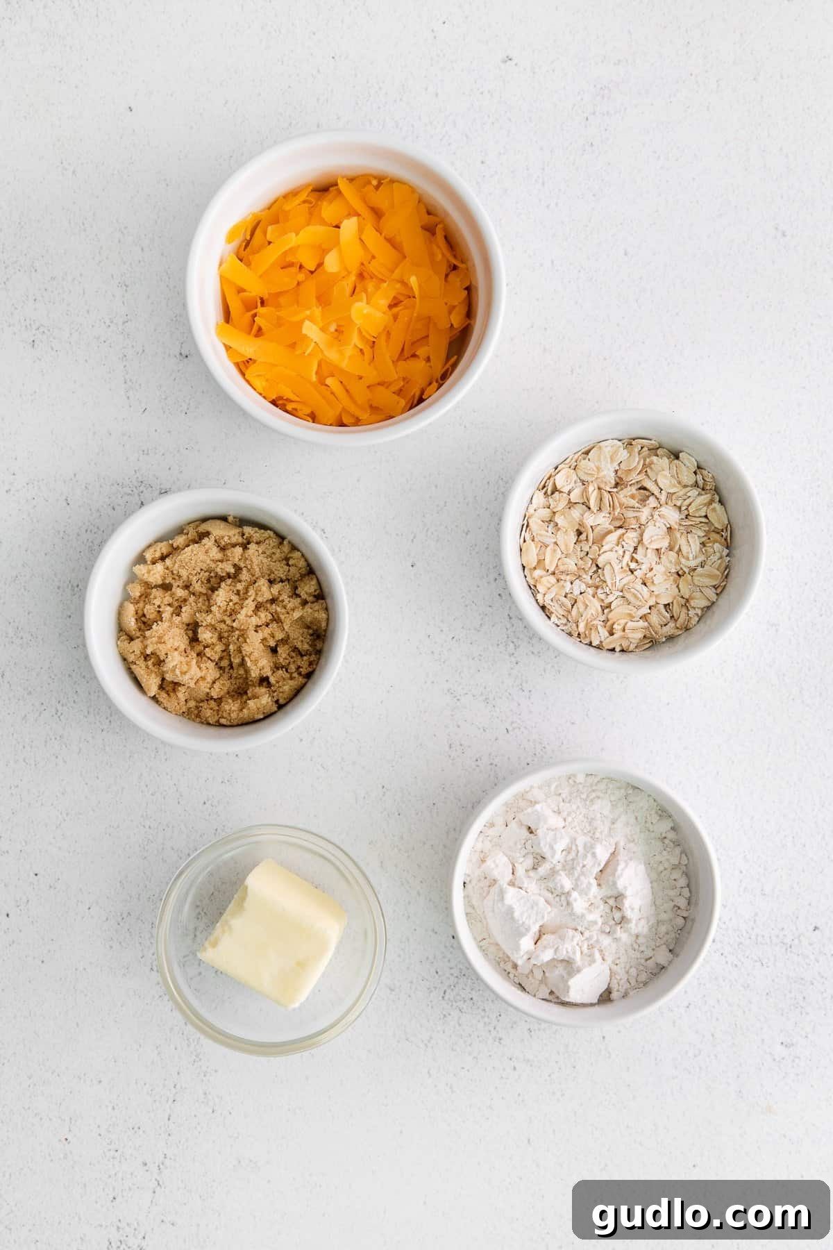 Bowls containing flour, rolled oats, brown sugar, shredded sharp cheddar cheese, and melted butter for the crumble topping.