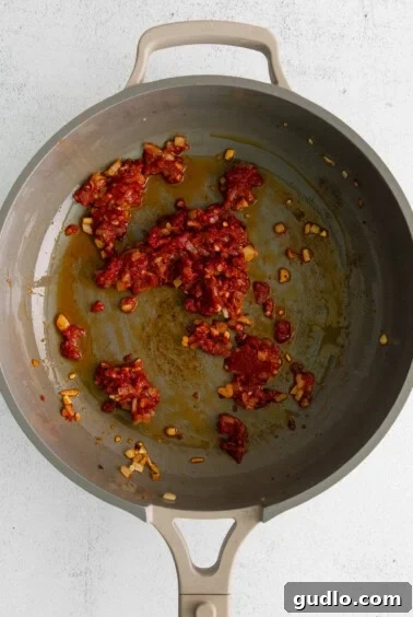 Tomato paste being added to a pan already containing olive oil, onions, and garlic, during the initial stages of making vodka sauce.