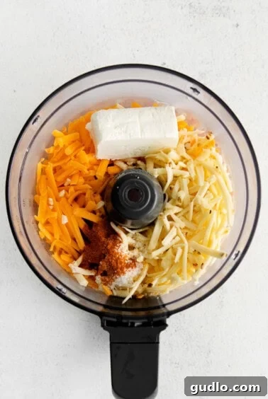 A food processor bowl with shredded cheeses, cream cheese, salt, and paprika, ready for blending.