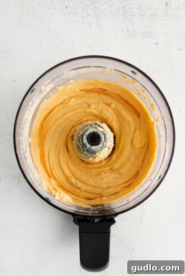 A food processor blending cheese ingredients into a thick, yellow batter, with a spatula nearby for scraping.