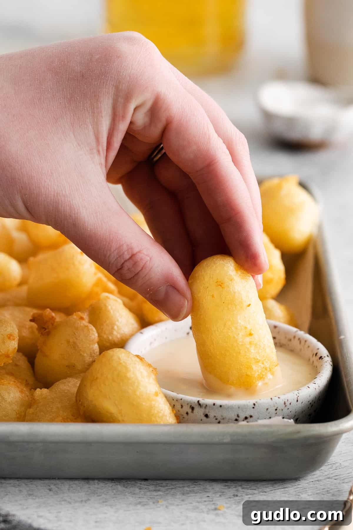 Dipping Fried Cheese Curds in Aioli A close-up shot of a golden-brown fried cheese curd being dipped into a small white bowl of creamy garlic aioli, highlighting its appetizing crispiness.