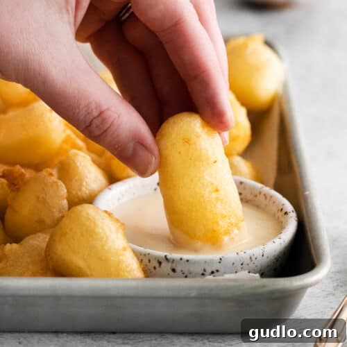 Artisan Fried Cheese Bites 2 A close-up of a person dipping a crispy fried cheese curd into a creamy white sauce, with more curds on a platter in the background.