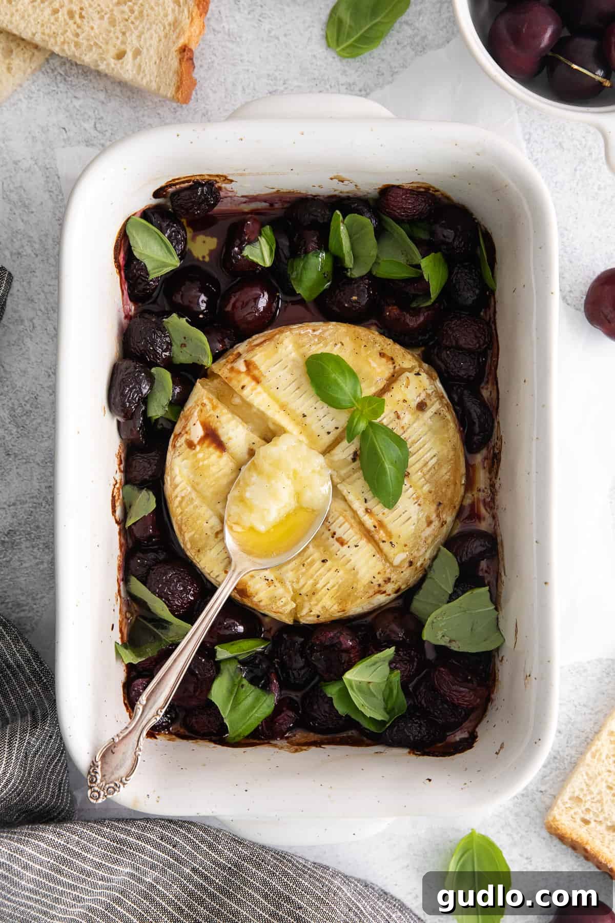 Grilled brie cheese appetizer in a baking dish, adorned with fresh cherries, balsamic glaze, and basil.
