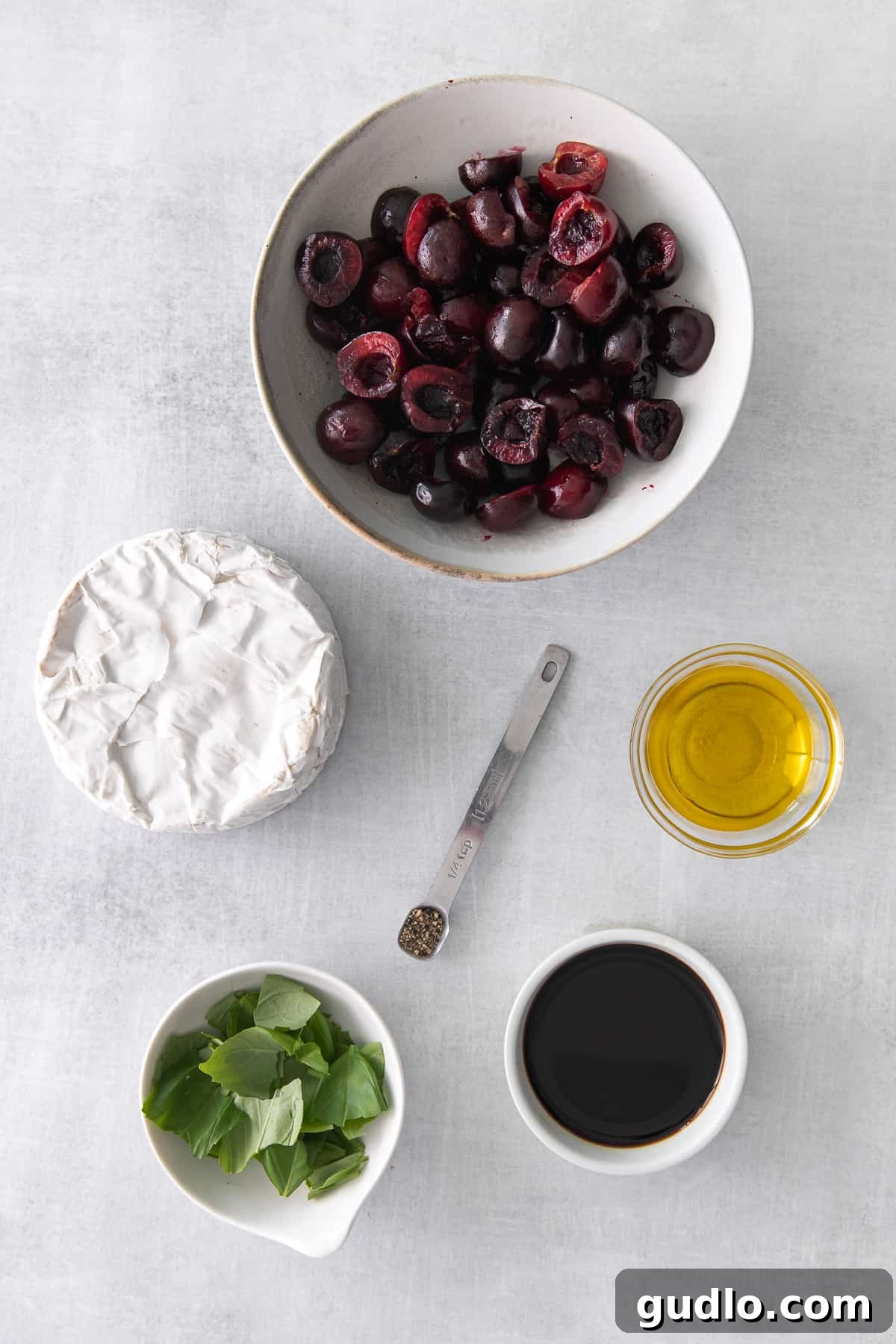 Ingredients for a grilled brie cheese appetizer displayed on a cutting board, including a brie wheel, fresh cherries, basil, balsamic vinegar, and olive oil.