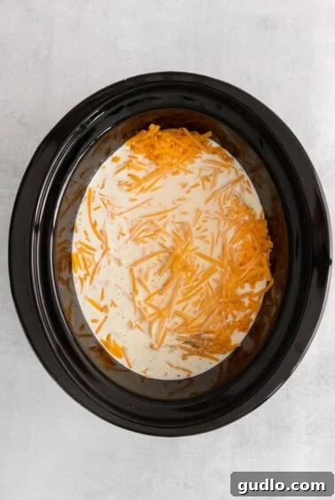 The creamy milk and cheese mixture being stirred with uncooked noodles in a slow cooker.