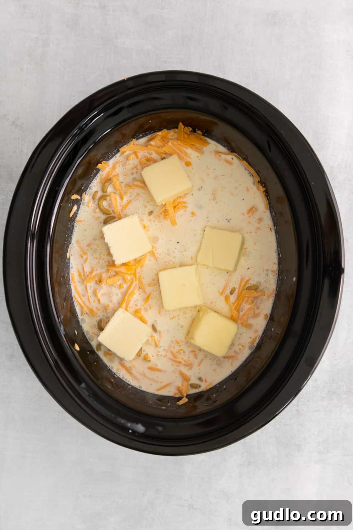 Butter Added to Crockpot Mac and Cheese Fresh pads of unsalted butter spread evenly over the mac and cheese mixture in a slow cooker before cooking, ready to melt into the creamy sauce.