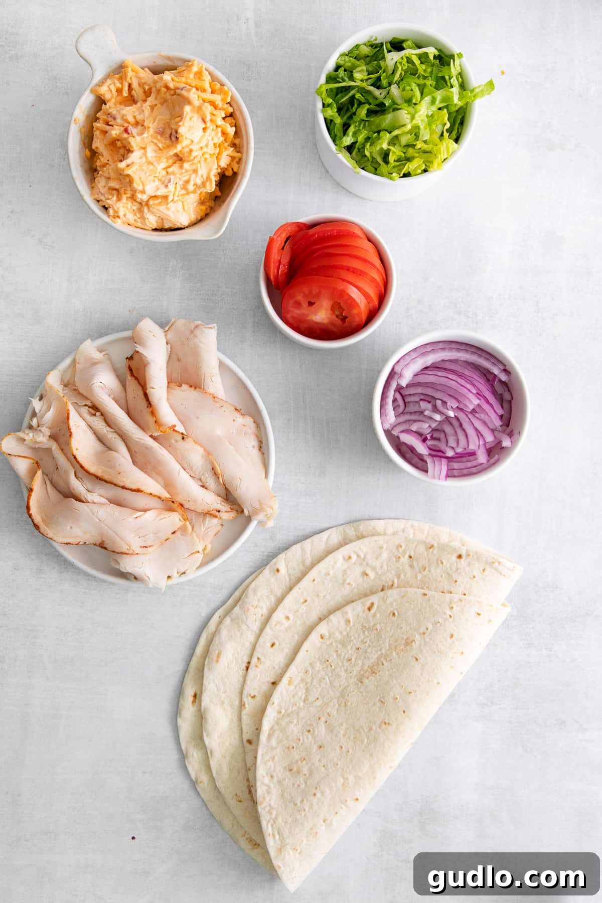 All the essential ingredients laid out on a wooden board: a bowl of pimento cheese, shredded lettuce, sliced tomato, sliced red onion, oven-roasted turkey slices, and soft flour tortillas.