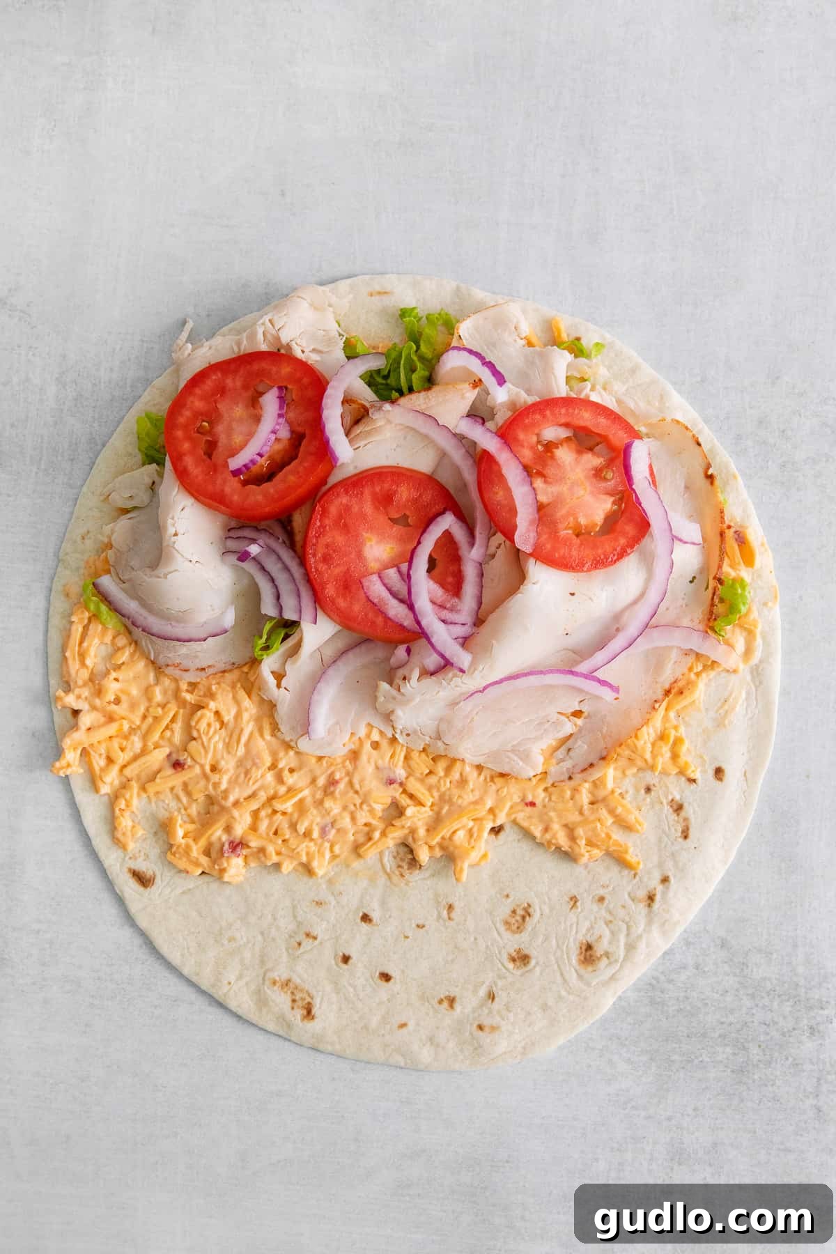 Layers of turkey, shredded lettuce, sliced tomato, and red onion atop pimento cheese on a tortilla, perfectly prepped for rolling.