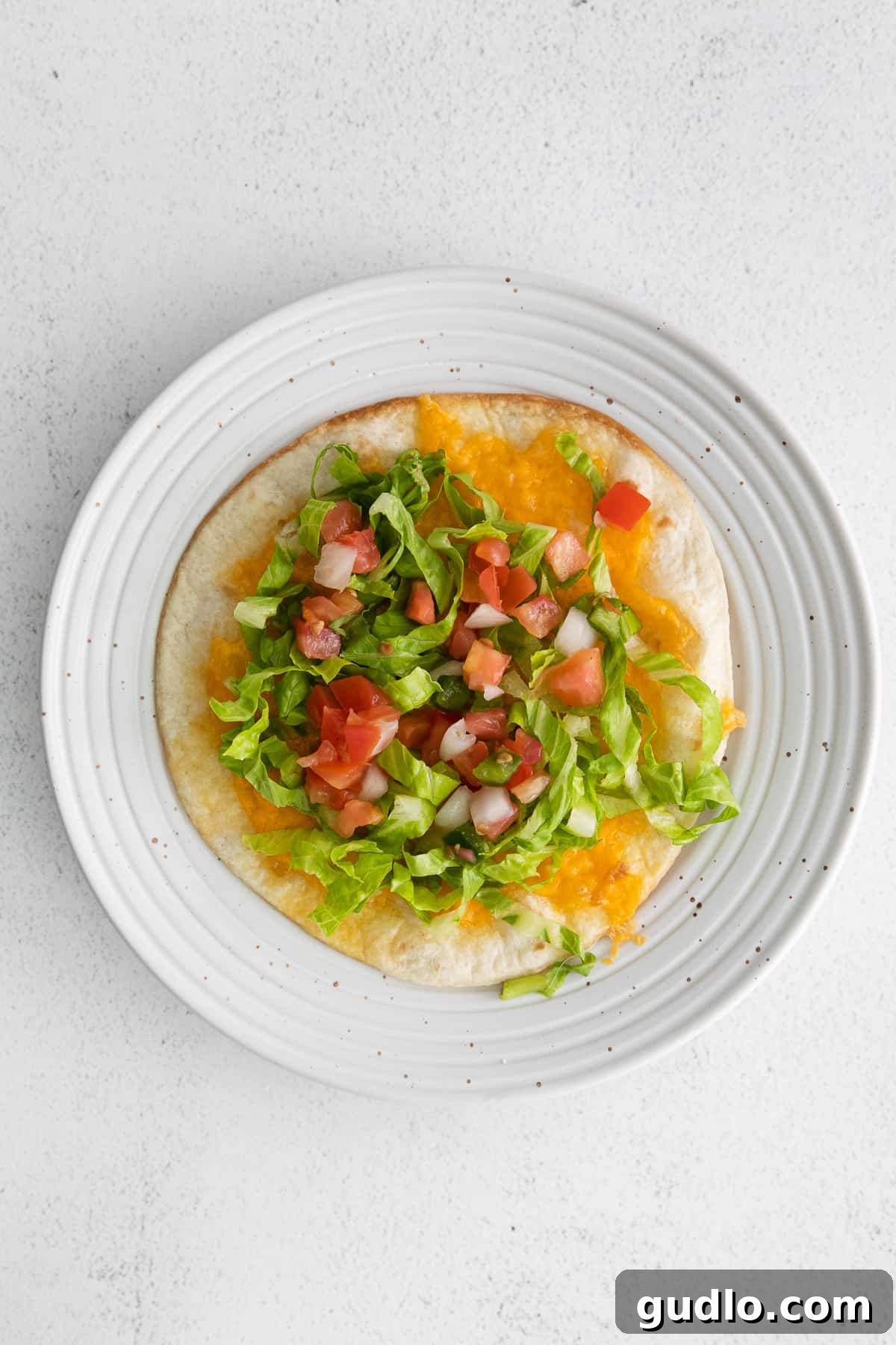 The initial layers of a copycat Applebee's quesadilla burger being built on a plate, featuring lettuce, pico de gallo, and a cheesy tortilla.