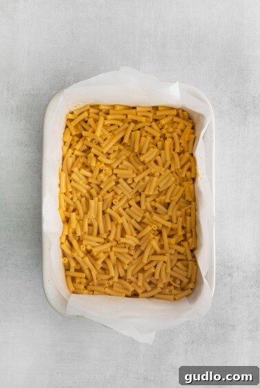 Creamy mac and cheese spread evenly in a casserole dish lined with parchment paper, chilling.