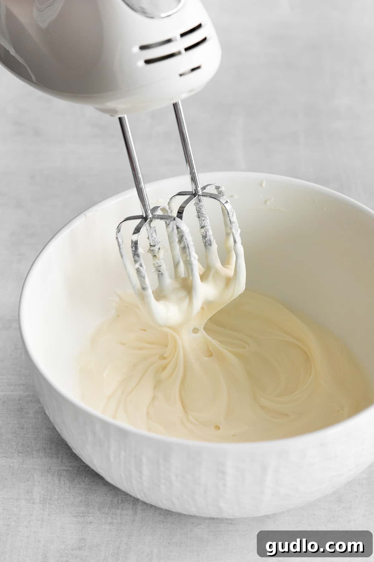 Homemade whipped cream cheese frosting being expertly mixed in a bowl with a hand mixer, highlighting its thick and creamy texture as it comes together.