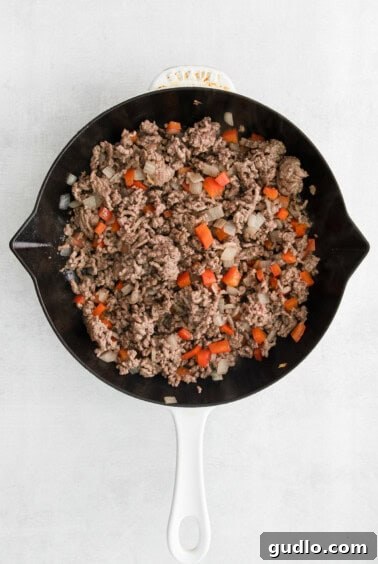A skillet filled with seasoned ground beef, diced red bell peppers, and onions, being sautéed on the stovetop.
