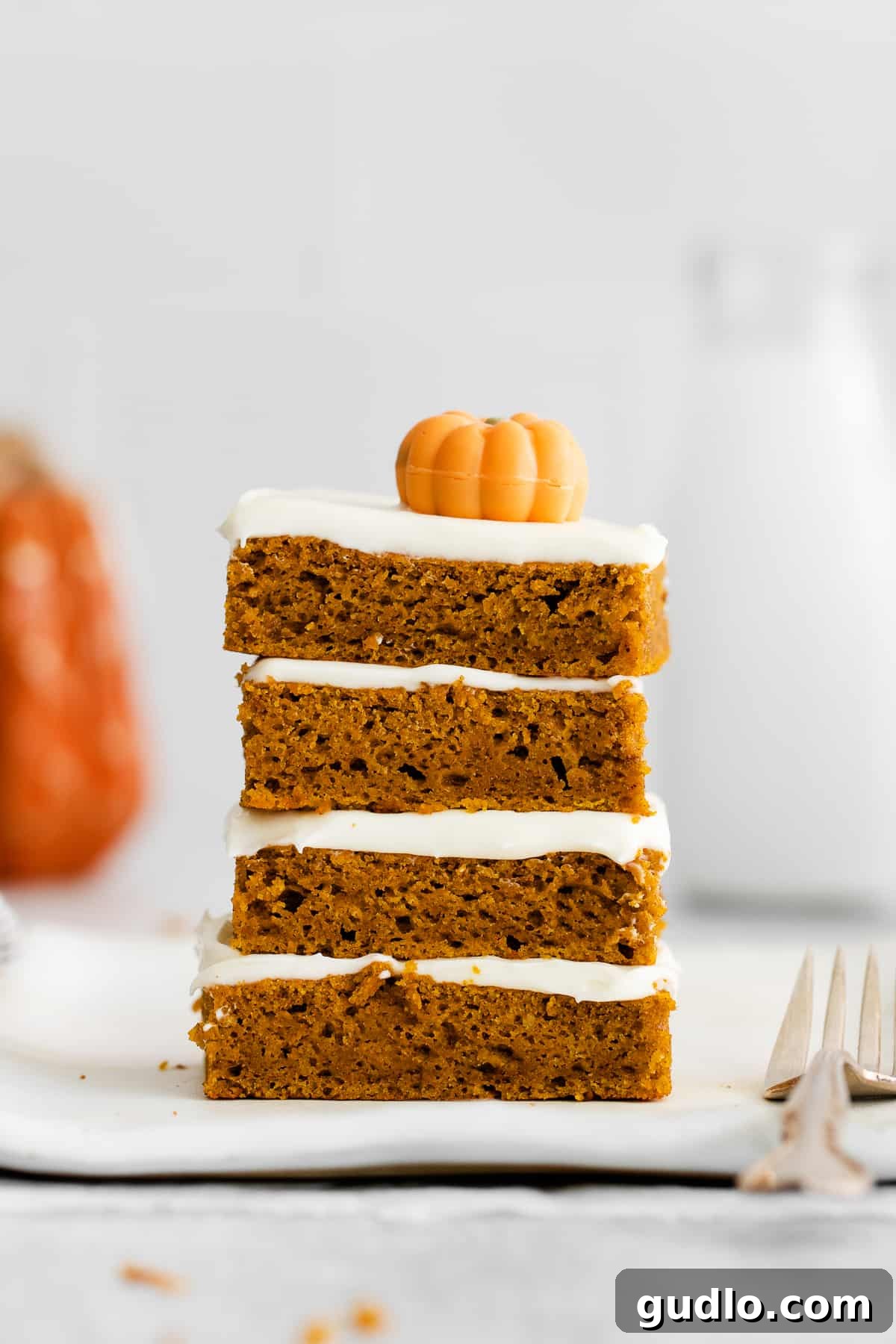 Four delicious homemade pumpkin bars, neatly stacked on a white plate, showcasing their fluffy frosting and inviting golden-orange hue, ready to be enjoyed as a perfect fall dessert.