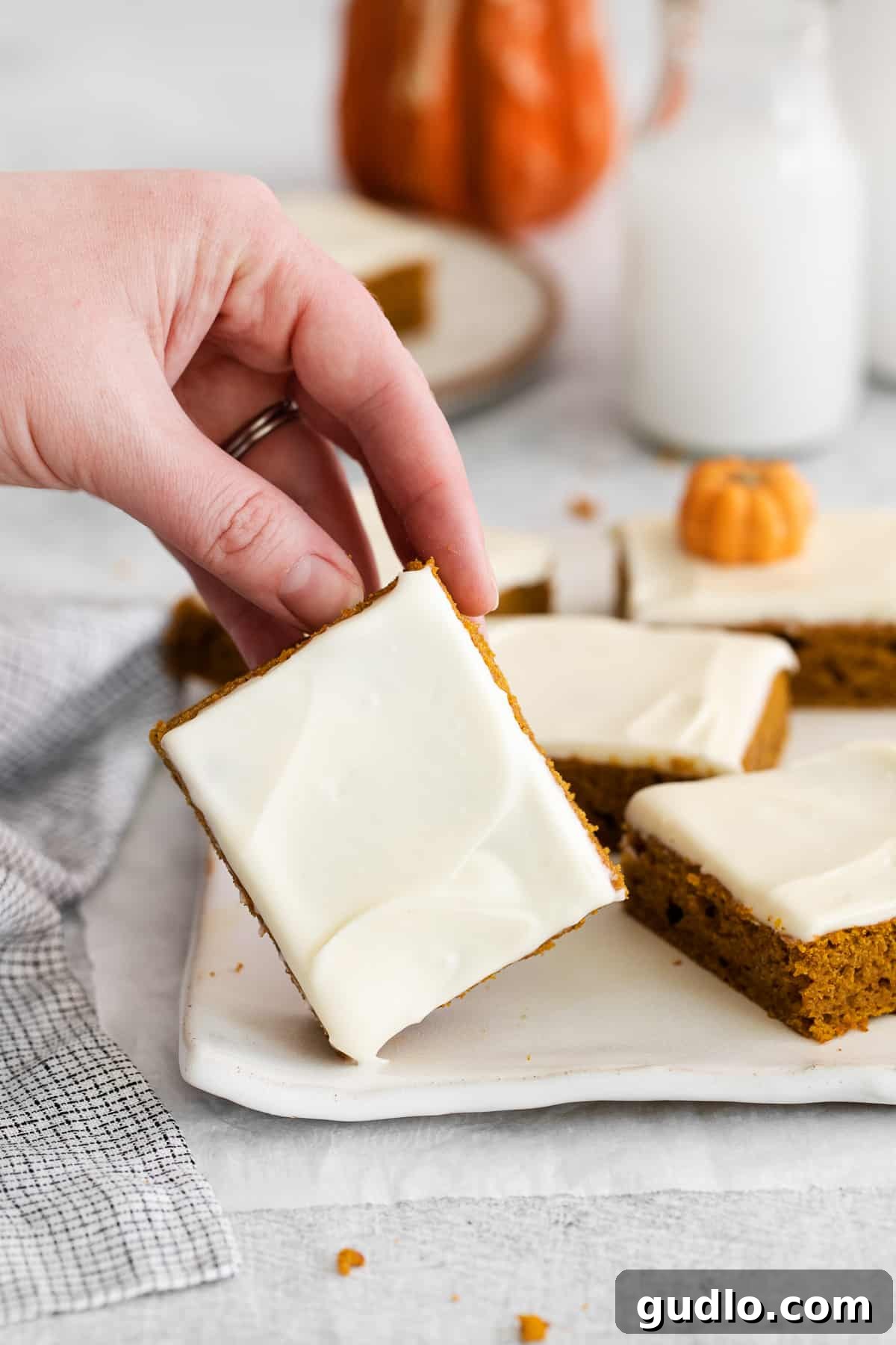 A hand gracefully holding a single frosted pumpkin bar on a small white plate, showcasing its soft texture and creamy topping.
